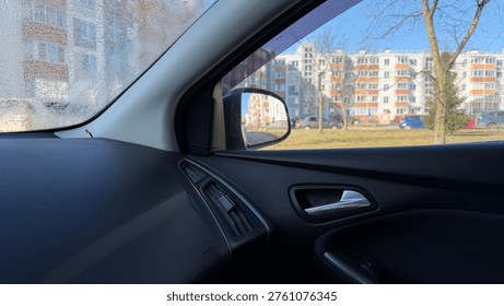 View from inside a car through a slightly open window. Black door panel, side mirror, and sunny residential buildings outside.