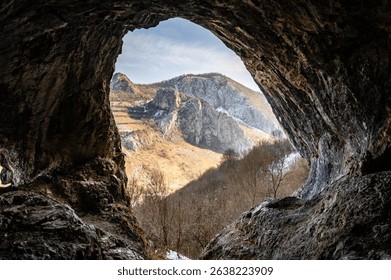 View from inside a cave looking out at rugged mountains under a bright sky, highlighting natural textures and formations.