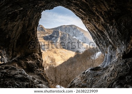 View from inside a cave looking out at rugged mountains under a bright sky, highlighting natural textures and formations.