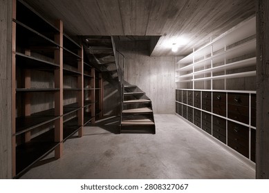 A view inside a concrete basement featuring extensive shelving units and a central staircase, suggesting a storage or utility space.