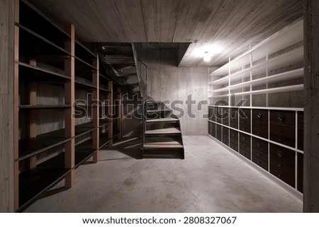 A view inside a concrete basement featuring extensive shelving units and a central staircase, suggesting a storage or utility space.