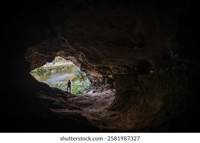 View from inside a dark cave looking out to a lush green landscape with a river and a person standing at the entrance.