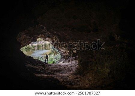 View from inside a dark cave looking out to a lush green landscape with a river and a person standing at the entrance.