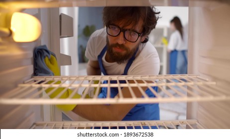 View From Inside Empty Refrigerator Of Man Wearing Rubber Gloves And Apron Cleaning Shelves. Male Janitor Washing Dirty Fridge With Sprayer And Cloth. House Cleaning Service Concept