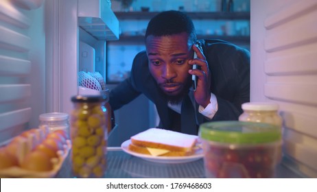 View Inside Fridge Of Tired Exhausted African Businessman In Suit Opens The Door Choosing What To Eat While Talking With Client By Smartphone