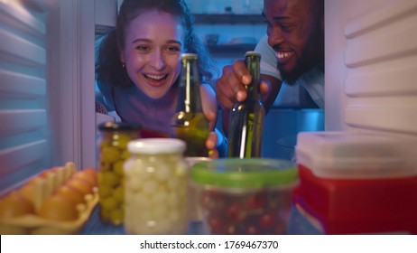 View Inside Fridge Of Young Multiethnic Couple Opening Refrigerator And Taking Cool Bottles Of Beer Holding Bowl With Popcorn Ready To Watch Movie Or Sport Game At Home