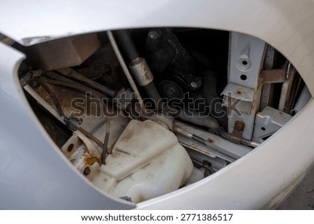 View inside headlight housing area of car with removed front lamp, showing fluid reservoir, hoses and structural elements behind bumper.