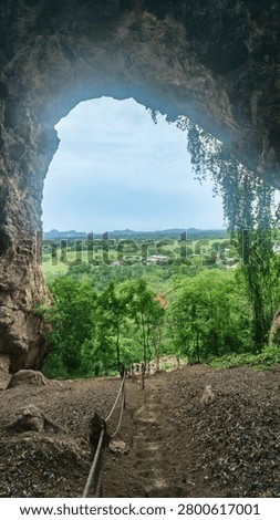 View from inside a limestone cave looking out through natural rock arch window opening to lush green tropical trees and blue sky, dramatic cave interior framing outdoor landscape, Thailand cave tourism photography