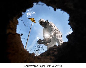 View from inside pit of ecologist digging pit by shovel, wearing gas mask and protective overalls outdoors, standing near warning sign with skull and crossbones, on background of blue cloudy sky.