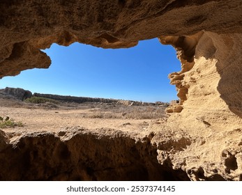 A view from inside a rocky cave, looking out over a sunny, open landscape with clear blue skies. The rugged, textured rock formations frame the natural scene beautifully. - Powered by Shutterstock