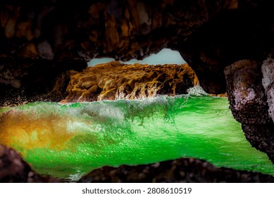 View from inside a rocky cave looking out at a powerful wave breaking in the bright green ocean water with sunlit cliffs beyond