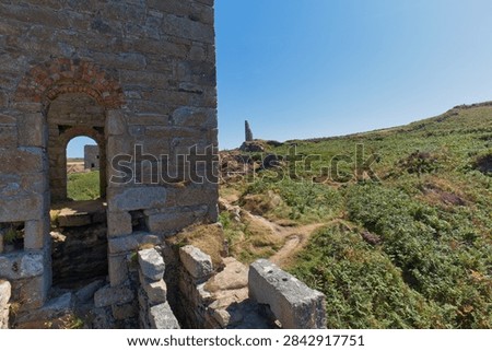 A view from inside a stone engine house ruin, part of Wheal Edward in Cornwall, looking out over a path through green heathland towards a distant mining structure.
