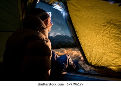 View From Inside A Tent On The Male Hiker Have A Rest In His Camping At Night. Man With A Headlamp Sitting In The Tent Near Campfire
