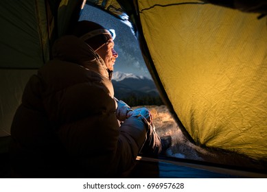 View From Inside A Tent On The Male Tourist Have A Rest In His Camping At Night. Man With A Headlamp Sitting In The Tent Near Campfire Under Beautiful Night Sky Full Of Stars And Milky Way