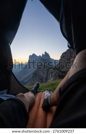 View from inside a tent, showing a person's legs resting on a mat, overlooking a stunning mountain landscape at sunrise or sunset.