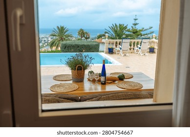 View from inside through a window onto a terrace dining table, swimming pool, and sea beyond