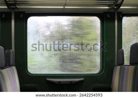 View from inside a train carriage showing empty seats and a large window. The window is slightly foggy with condensation and displays a blurred view of green trees speeding by, evoking a sense of quie