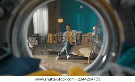 View from inside the washing machine, young man sitting on the couch in the room and smiles