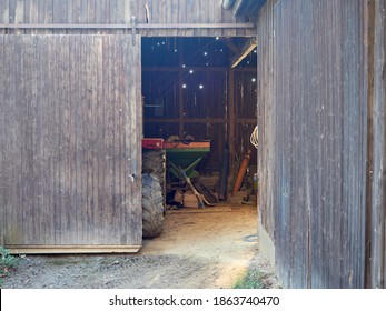 View Inside The Wooden Barn Through The Open Door. Old Barn With A Tractor Inside. Sun Rays Falling Into A Country House Through Holes In The Wall