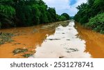 View of irrigation canal with weeds and debris flowing together with strong flowing dirty brown water due to soil and mud from forest after heavy rains and floods. La Nina crisis, Water pollution.