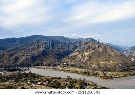 View from the Jvari Monastery to buildings of Mtskheta city surrounded by dense trees near the riverbank of winding Kura river flowing through lush, green hills and mountains. Georgia