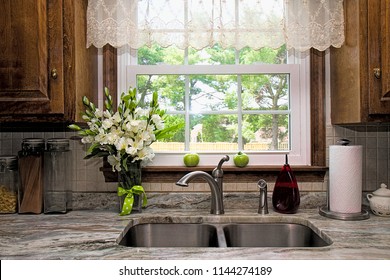 View of Kitchen sink, window view of backyard, curtains and flowers.  Two green tomatoes on window seal witing to ripe.  Beautiful designer gray/white/green granite countertop.