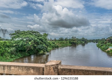 View Of The Kwanza (Cuanza) River, Tropical Vegetation On Banks, Electric Power Lines, Cloudy Sky As Background, In Angola