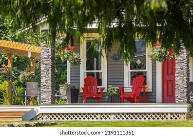 View of a large front porch with furniture and potted plants. Rustic, country style front porch seating with red Adirondack chairs and fresh flowers. Nobody, - Shutterstock ID 2191111351