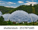 A view of large, geodesic domes made of glass and metal, surrounded by lush greenery and hills under a blue sky with clouds. At Eden Project, Cornwall, UK