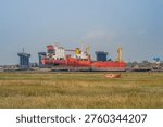 View of a large red ship partially dismantled, stranded in the mud of the Chittagong coast. In the foreground, a field of dry grass and a small orange rowboat contrast with the massive, rusted industrial structures of the shipbreaking yard.
