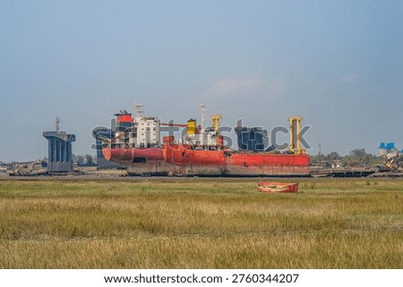 View of a large red ship partially dismantled, stranded in the mud of the Chittagong coast. In the foreground, a field of dry grass and a small orange rowboat contrast with the massive, rusted industrial structures of the shipbreaking yard.