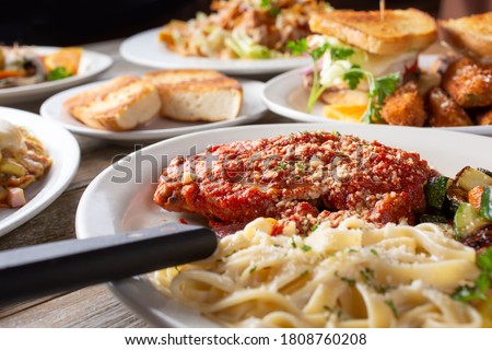 A view of a layout of several prepared entrees, featuring a plate of chicken parmigiana and pasta.