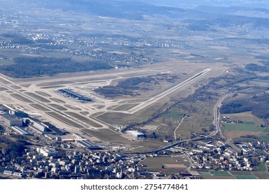View from the left airplane window after takeoff from Zurich Airport, showing the full runway layout from above on a clear sunny day.