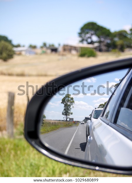 View from the left side rear view mirror of a car\
driving in a rural road with dry grass and farm fence on side. Cars\
following behind on a country road in remote area on a hot summer\
day.