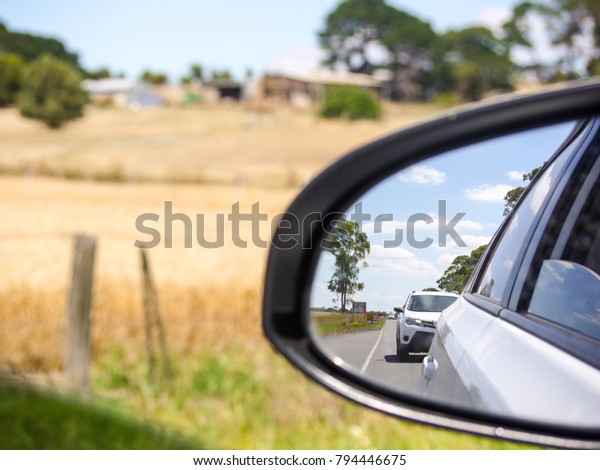 View from the left side rear view mirror of a car\
driving in a rural road with dry grass and farm fence on side. Cars\
following behind on a country road in remote area on a hot summer\
day.
