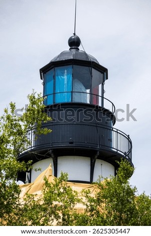 View of lighthouse through green leafy trees.  Pale sky, green leaves and lighthouse structure with tarp wrapped around.  Amelia Island lighthouse with renovation tarp.