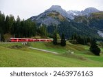 View of a local train traveling through a grassy meadow on the hillside with majestic alpine mountains in the background, in Bergun Village between Chur and St. Moritz, in Grisons, Switzerland