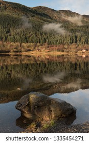 View Of The Loch Lomond Under A Blue Sky With A Big Rock Lying In The Water On The Foreground And Trees Reflected On The Mirror-like Lake As The Low Clouds Dissipate In The Trossachs National Park, UK