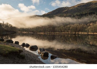 View Of The Loch Lomond Under A Blue Sky In A Misty Morning With A Line Of Stones And Trees Reflected On The Lake's Calm Water As The Low Clouds Pass Through In  The Trossachs National Park, Scotland,