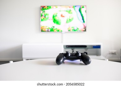 A View Of A Logo Free, Black Videogame Controller On A White Table. Tv Is In The Background.