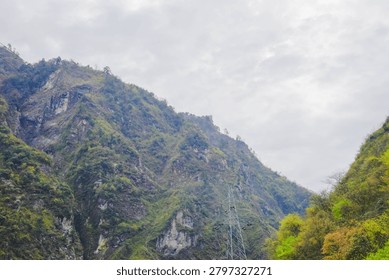 A view looking across a deep mountain valley, framed by lush green hillsides on the right and steep rocky cliffs on the left. An electrical transmission tower stands at the base of the gorge against a hazy sky.