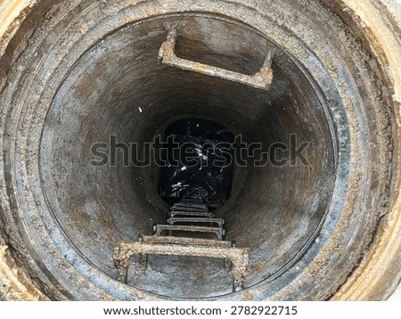 View looking down inside a deep underground manhole shaft with metal ladder rungs and sewer or utility access below.