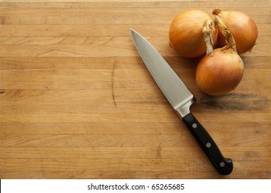 A view looking down on a group of onions and a large knife on a worn butcher block cutting board