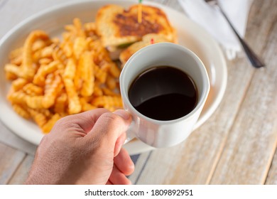 A View Looking Down Onto A Hand Holding A Mug Of Coffee, Over A Plate Of Food, In A Restaurant Or Kitchen Setting.