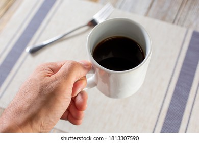 A View Looking Down Onto A Hand Holding A Mug Of Coffee, Over A Plate Setting, In A Restaurant Or Kitchen Setting.