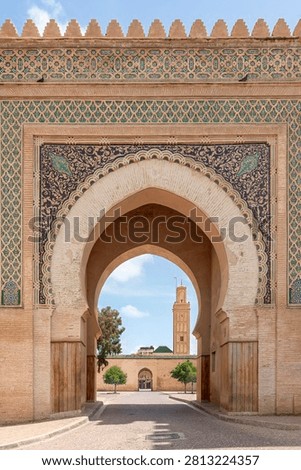 A view looking through the decorated arch of Bab Berdaine, its ornate zellige tilework frieze framing a perfect view of the tall minaret of the Mosque Berdaine rising beyond the gate.