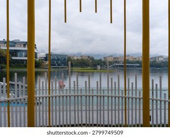 A view looking through the vertical yellow bars of an architectural pavilion towards a wide lake and distant mountains under cloudy skies. The scene represents perspective, architectural framing, peaceful lakeside atmosphere, and urban tourism.