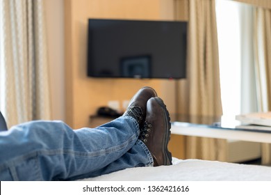 View Of A Man's Legs And Feet Relaxing On A Motel Bed In Front Of The TV. Man Is Dressed In Worn Jeans And Work Boots.