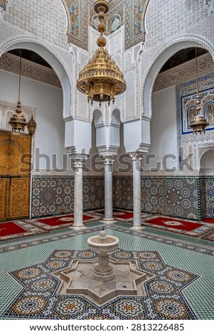 A view of the mausoleum interior shows multiple marble columns, elaborate chandeliers hanging from the painted ceiling, red prayer rugs on the floor, and carved stucco arches beyond a zellige fountain.