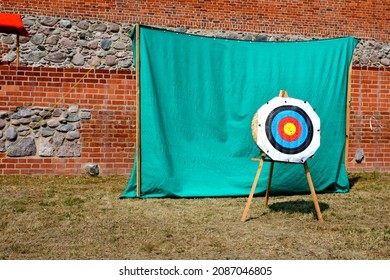 A view of a medieval shooting tange with a target in the shape of a round circle standing next to a green cloth situated next to the wall of a medieval castle or church seen on a sunny summer day